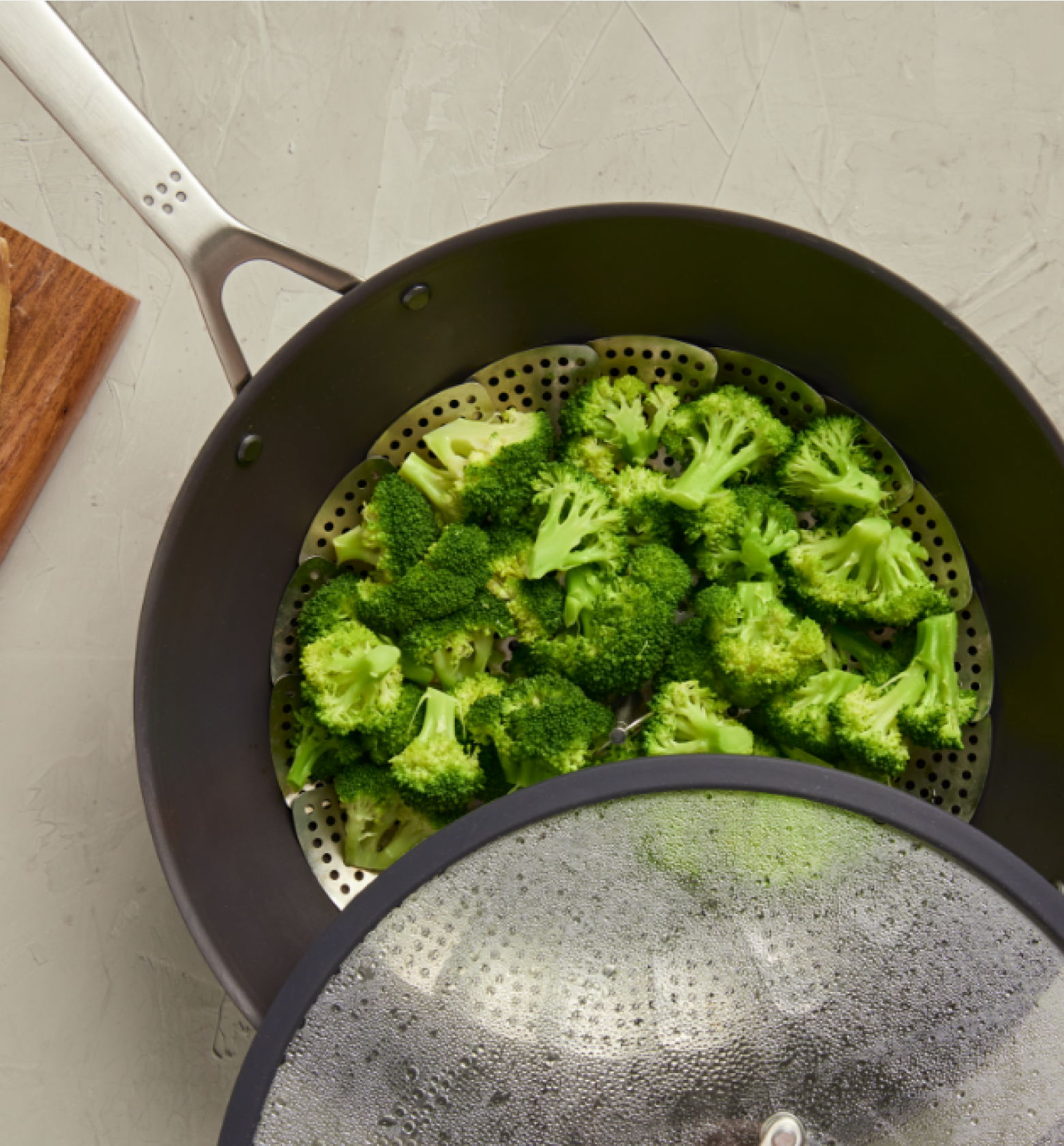 Fresh broccoli florets steaming in a metal steamer basket inside a dark nonstick pan